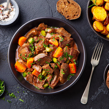 Beef meat and vegetables stew in black bowl with roasted baby potatoes. Dark background. Top view.